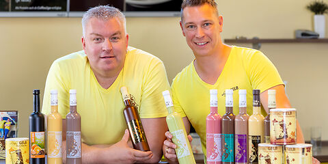 Zwei Besitzer der Manufaktur sitzen am Tisch und präsentieren zehn verschiedene Sorten Eierlikör. Beide tragen gelbe T-Shirts.
