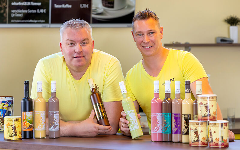 Zwei Besitzer der Manufaktur sitzen am Tisch und präsentieren zehn verschiedene Sorten Eierlikör. Beide tragen gelbe T-Shirts.