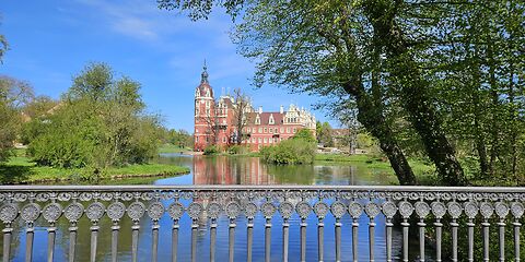 Das Schloss in Bad Muskau ist hinter dem dekorativen Geländer einer Brücke zu sehen. Vor dem Schloss liegt ein kleiner See, der auf beiden Seiten von üppigem Grün umgeben ist. Das Blau der Wasseroberfläche geht harmonisch in das Blau des Himmels über.