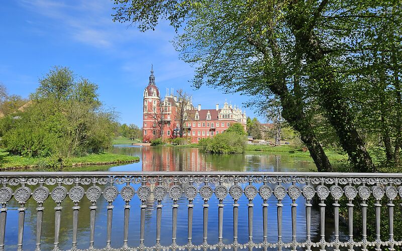 Das Schloss in Bad Muskau ist hinter dem dekorativen Geländer einer Brücke zu sehen. Vor dem Schloss liegt ein kleiner See, der auf beiden Seiten von üppigem Grün umgeben ist. Das Blau der Wasseroberfläche geht harmonisch in das Blau des Himmels über.