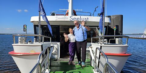 Die Bordcrew des Ausflugsschiffes „Bärwalder Seelust“ winkt den Gästen dankend zu. Das weiße Schiff ist aus nächster Nähe von hinten zu sehen. Im Vordergrund befindet sich die bewegliche Einstiegsplattform des Schiffes.