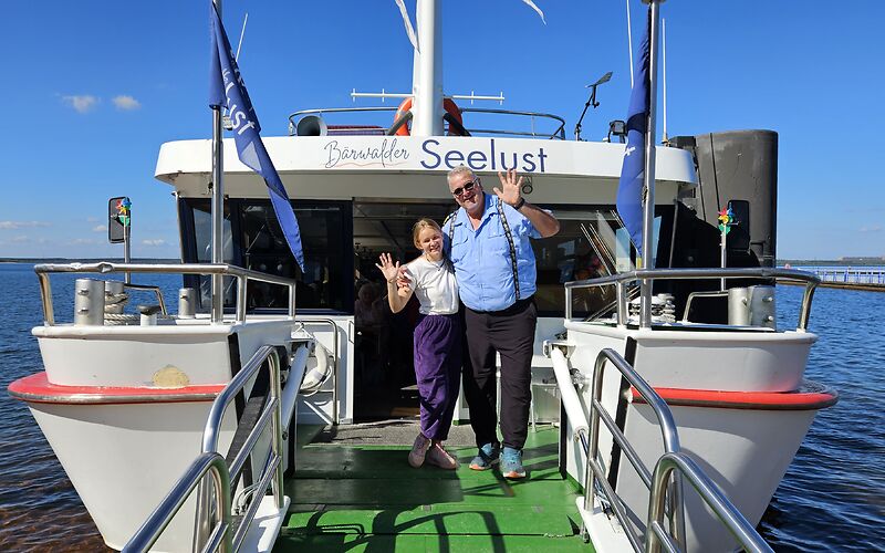 Die Bordcrew des Ausflugsschiffes „Bärwalder Seelust“ winkt den Gästen dankend zu. Das weiße Schiff ist aus nächster Nähe von hinten zu sehen. Im Vordergrund befindet sich die bewegliche Einstiegsplattform des Schiffes.