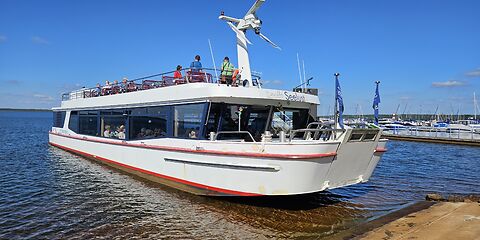 Ein weißes Ausflugsschiff mit Touristen auf dem Bärwalder See an einem sonnigen Tag. Der blaue Himmel und das klare, blaue Wasser prägen die Szenerie. Auf der rechten Seite sind Motorboote und ein Steg zu sehen.