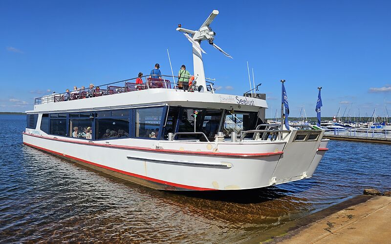 Ein weißes Ausflugsschiff mit Touristen auf dem Bärwalder See an einem sonnigen Tag. Der blaue Himmel und das klare, blaue Wasser prägen die Szenerie. Auf der rechten Seite sind Motorboote und ein Steg zu sehen.