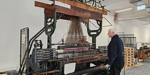 Zwei Techniker arbeiten an einem historischen Jacquard-Kurbelwebstuhl im Forster Museum. Sie stehen auf gegenüberliegenden Seiten der großen Maschine und prüfen deren Mechanik, um den Webstuhl wieder in Betrieb zu nehmen. Der Webstuhl besteht aus zahlreichen Metall- und Holzteilen sowie dem für die Jacquard-Technik typischen Aufsatz mit den Lochkarten.