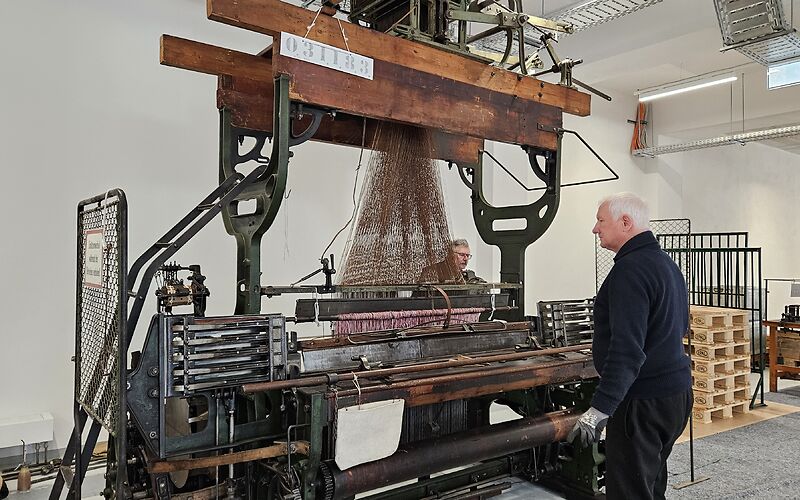 Zwei Techniker arbeiten an einem historischen Jacquard-Kurbelwebstuhl im Forster Museum. Sie stehen auf gegenüberliegenden Seiten der großen Maschine und prüfen deren Mechanik, um den Webstuhl wieder in Betrieb zu nehmen. Der Webstuhl besteht aus zahlreichen Metall- und Holzteilen sowie dem für die Jacquard-Technik typischen Aufsatz mit den Lochkarten.