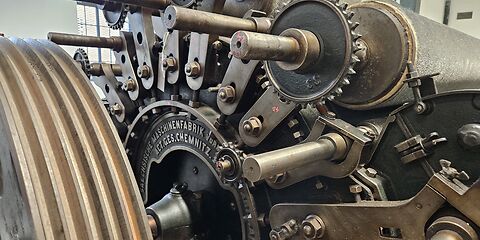 Das Foto zeigt eine große Nahaufnahme eines Maschinenteils einer Textilmaschine. Zu erkennen sind zahlreiche Schrauben und Zahnräder sowie die eingravierte Aufschrift „Sächsische Maschinenfabrik Vorwerk ACT. GES. Chemnitz“.