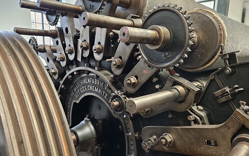 Das Foto zeigt eine große Nahaufnahme eines Maschinenteils einer Textilmaschine. Zu erkennen sind zahlreiche Schrauben und Zahnräder sowie die eingravierte Aufschrift „Sächsische Maschinenfabrik Vorwerk ACT. GES. Chemnitz“.