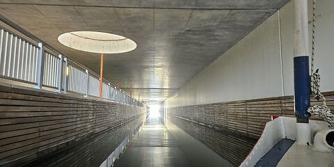 Auf dem Foto ist ein moderner Tunnel zwischen dem Großräschener See und dem Sedlitzer See zu sehen. Durch runde Öffnungen in der Decke fällt Tageslicht in den Tunnel. Auf der Wasseroberfläche ist eine schöne Spiegelung des Lichts zu sehen.