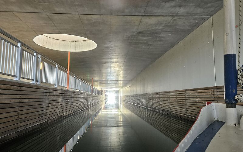 Auf dem Foto ist ein moderner Tunnel zwischen dem Großräschener See und dem Sedlitzer See zu sehen. Durch runde Öffnungen in der Decke fällt Tageslicht in den Tunnel. Auf der Wasseroberfläche ist eine schöne Spiegelung des Lichts zu sehen.