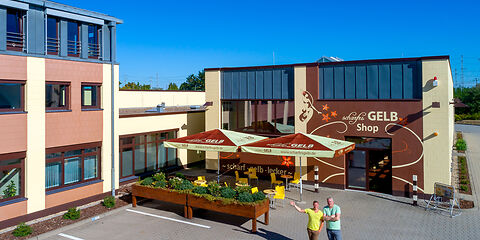 Der Haupteingang der Manufaktur scharfesGELB von außen. Die Fassade ist in Gelb, Braun und Schwarz gestaltet. Zwei Besitzer stehen am Eingang und winken freundlich.