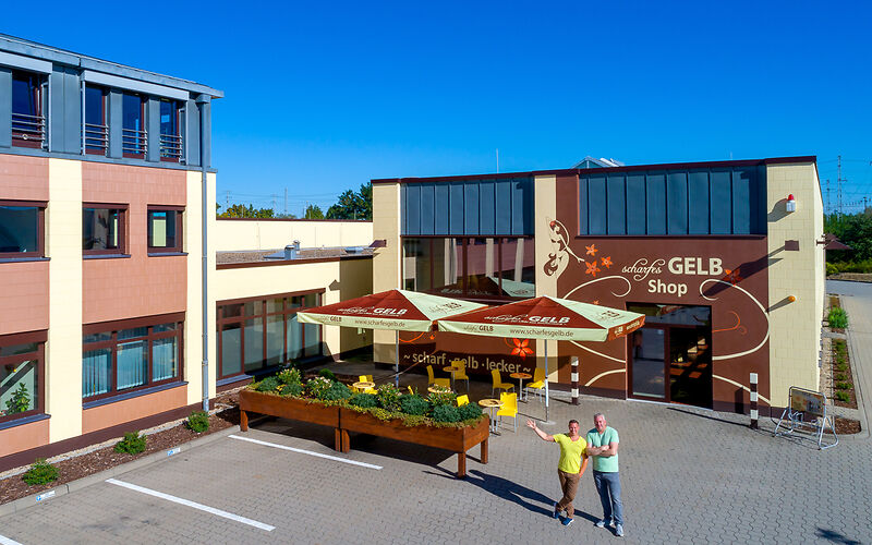 Der Haupteingang der Manufaktur scharfesGELB von außen. Die Fassade ist in Gelb, Braun und Schwarz gestaltet. Zwei Besitzer stehen am Eingang und winken freundlich.