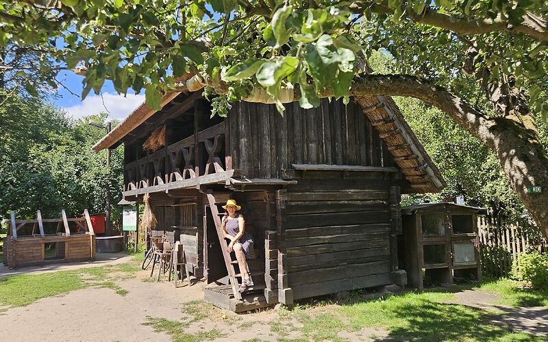 Auf dem Foto ist ein typisches hölzernes Wirtschaftsgebäude im Freilandmuseum in Lehde mit einem Reetdach zu sehen. Das Gebäude steht neben einem großen Baum. Auf der schmalen Holztreppe des Hauses sitzt eine Touristin mit einem gelben Hut.