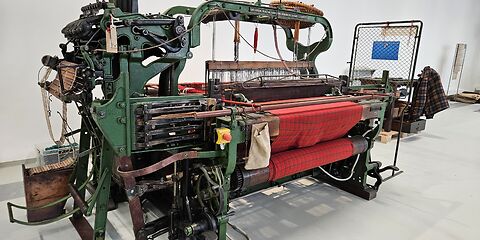 Zu sehen ist ein historischer Kurbel-Buckskin-Webstuhl, der grün lackiert ist. Auf der linken Seite der Maschine befinden sich die für den Webprozess benötigten Lochkarten. Im Webstuhl ist ein rot karierter Stoff eingespannt, dessen Muster bereits deutlich erkennbar ist.