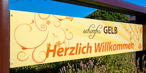 Gelbes Werbeschild mit der Aufschrift "scharfesGELB" in Braun und ‚Herzlich Willkommen‘ in Orange.