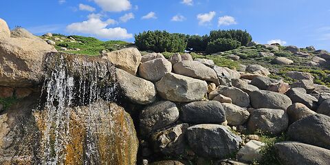 Große Steine umrahmen einen kleinen Wasserfall im Findlingspark Nochten. Der blaue Himmel mit einigen weißen Wölkchen verleiht der Aufnahme eine freundliche und idyllische Atmosphäre.