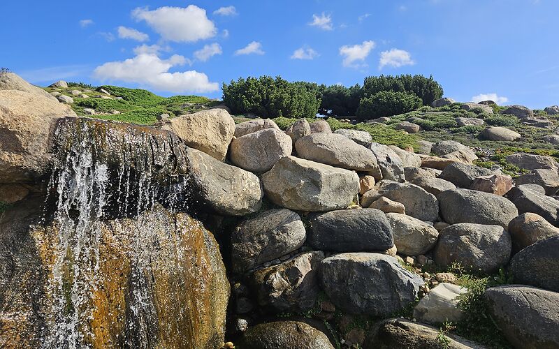 Große Steine umrahmen einen kleinen Wasserfall im Findlingspark Nochten. Der blaue Himmel mit einigen weißen Wölkchen verleiht der Aufnahme eine freundliche und idyllische Atmosphäre.