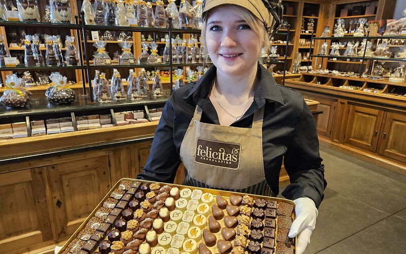 Im SchokoLaden der Confiserie Felicitas in Hornow präsentiert eine Frau auf einem Tablet eine Auswahl verschiedener Pralinen. Im Hintergrund sind mehrere Figuren aus Schokolade zu sehen.