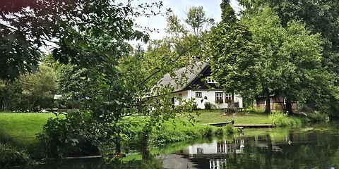 Blick vom Spreekanal auf ein traditionelles Spreewaldhaus, das von einem schönen, grünen Grundstück umgeben ist.