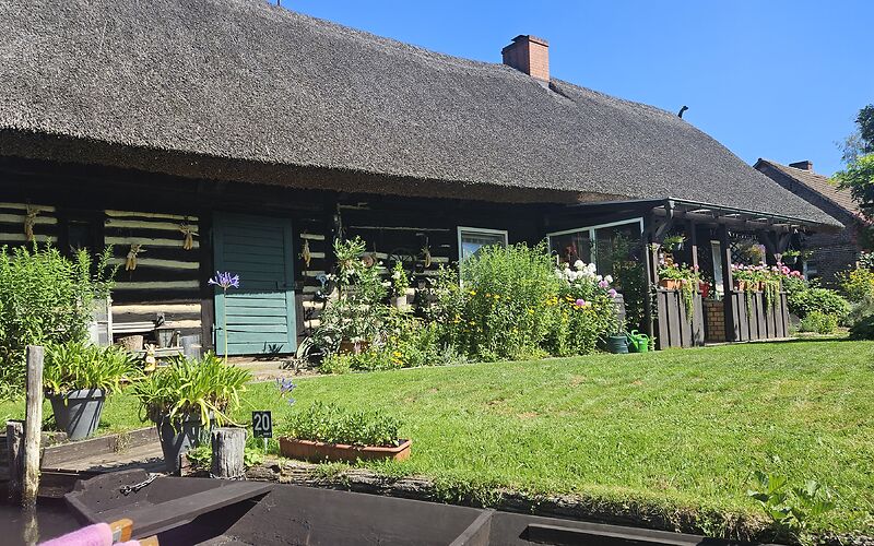 Zu sehen ist ein typisches Spreewaldhaus mit einem Reetdach. Direkt am Haus wachsen dekorative Blumen. Vor dem Haus liegt eine grüne Wiese. Am Ufer liegt ein traditioneller Holzkahn.