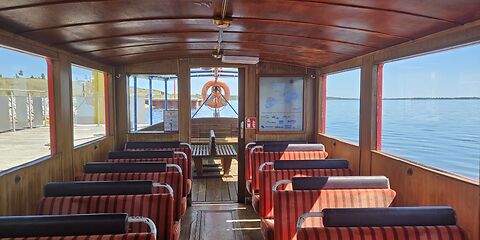 Im Inneren des Schiffs „Wilde Ilse“ befinden sich links und rechts rote Sitzbänke. Durch die großen Fenster bietet sich ein weiter Panoramablick auf den See. Im Hintergrund ist ein Rettungsring zu sehen.