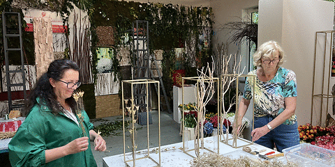 Zwei Frauen arbeiten an einem Tisch mit Trockenblumen und Holzrahmen in einem Raum mit Pflanzenwänden und Holzbalkendecke