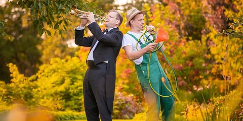 Auf dem Foto ist ein musikalisches Duo mit dem Namen „Aufgeweckte Gartenklänge“ zu sehen. Links steht ein Musiker aus einem Sinfonieorchester. Er trägt einen schwarzen Smoking mit weißem Hemd und spielt eine Trompete. Rechts steht ein Gärtner in Arbeitskleidung. Er hält einen grünen Gartenschlauch und benutzt ihn wie ein Musikinstrument, indem er hineinbläst.  Die beiden Personen stehen in einem Park. Im Hintergrund sind Bäume mit herbstlich gefärbten Blättern in Gelb-, Orange- und Rottönen zu sehen. Die Szene wirkt heiter, kreativ und leicht humorvoll.