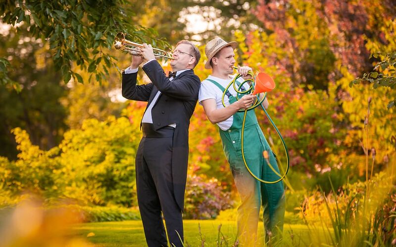 Auf dem Foto ist ein musikalisches Duo mit dem Namen „Aufgeweckte Gartenklänge“ zu sehen. Links steht ein Musiker aus einem Sinfonieorchester. Er trägt einen schwarzen Smoking mit weißem Hemd und spielt eine Trompete. Rechts steht ein Gärtner in Arbeitskleidung. Er hält einen grünen Gartenschlauch und benutzt ihn wie ein Musikinstrument, indem er hineinbläst.  Die beiden Personen stehen in einem Park. Im Hintergrund sind Bäume mit herbstlich gefärbten Blättern in Gelb-, Orange- und Rottönen zu sehen. Die Szene wirkt heiter, kreativ und leicht humorvoll.