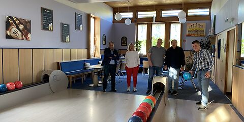 Die Gruppe steht in einem hellen Bowlingraum, eine Person wirft eine blaue Kugel auf der Bahn