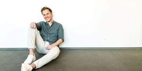 Junger Mann sitzt locker auf einem dunklen Holzfußboden und einer weißen Wand.