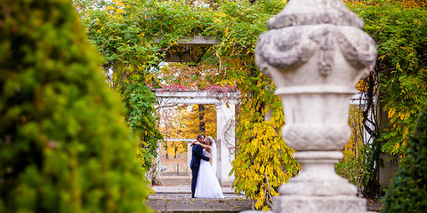 Rechts steht eine Amphore aus Ton im Hintergrund umarmend und küssend die Braut in einem weißen Kleid und der Bräutigam im blauen Anzugdie Bräutogam