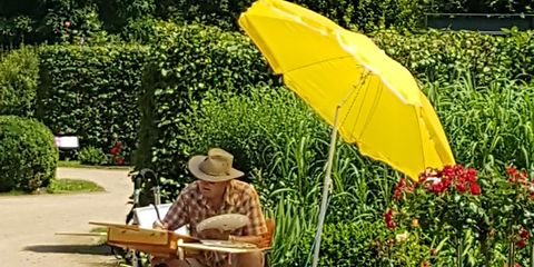ein Mann mit Strohhut sitzt unter einen gelben Sonnenschirm am Wegrand und zeichnet auf einem provisorischen Tisch