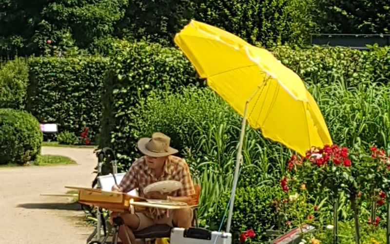 ein Mann mit Strohhut sitzt unter einen gelben Sonnenschirm am Wegrand und zeichnet auf einem provisorischen Tisch
