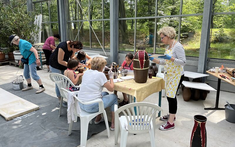 An einem Tisch sitzen mehrere Frauen, die Vasen dekorieren. Im Hintergrund sind durch die Fensterwand des Gebäudes die Bäume im Rosengarten zu sehen.