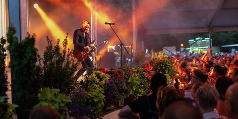 Auf dem Foto sieht man ein Konzert im Rosengarten Forst. Viele Menschen stehen vor der Bühne. Sie lachen und freuen sich. Die Bühne und das Publikum sind bunt beleuchtet. Es herscht eine tolle Atmosphäre. 