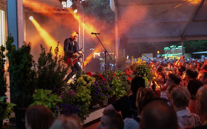 Auf dem Foto sieht man ein Konzert im Rosengarten Forst. Viele Menschen stehen vor der Bühne. Sie lachen und freuen sich. Die Bühne und das Publikum sind bunt beleuchtet. Es herscht eine tolle Atmosphäre. 