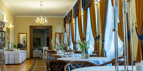 Auf dem Foto ist das prachtvolle Restaurant im Pałac Brody zu sehen. Große Schlossgardinen, runde Tische mit weißen Tischdecken und historische Lampen verleihen dem Raum eine elegante, historische Atmosphäre.