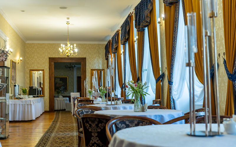 Auf dem Foto ist das prachtvolle Restaurant im Pałac Brody zu sehen. Große Schlossgardinen, runde Tische mit weißen Tischdecken und historische Lampen verleihen dem Raum eine elegante, historische Atmosphäre.