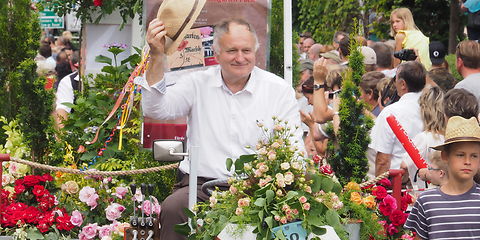 Ein Mann mit einem weißen Hemd sitzt auf einem Trakor und hält einen Strohhut in der rechten Hand und grüßt die Zuschauer am Straßenrand. Im Hintergrund ein Anhänger geschmückt mit bunten Blumen