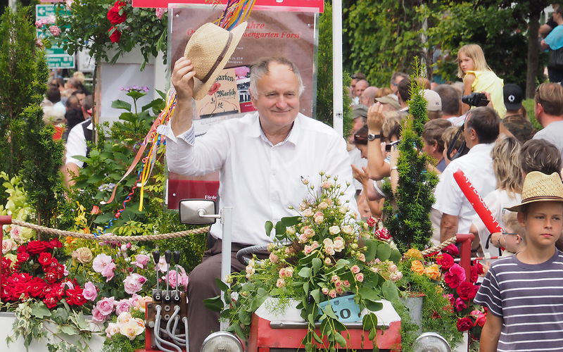 Ein Mann mit einem weißen Hemd sitzt auf einem Trakor und hält einen Strohhut in der rechten Hand und grüßt die Zuschauer am Straßenrand. Im Hintergrund ein Anhänger geschmückt mit bunten Blumen
