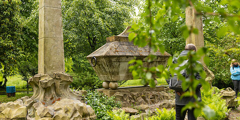 Im Schlosspark Brody ist ein historischer steinerner Sarkophag zu sehen, der von zwei hohen Obelisken eingerahmt wird. Das Denkmal steht inmitten einer dicht begrünten Parkanlage mit Farnen und Bäumen. Im Vordergrund und seitlich sind einige Besucherinnen und Besucher zu sehen.