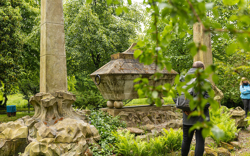 Im Schlosspark Brody ist ein historischer steinerner Sarkophag zu sehen, der von zwei hohen Obelisken eingerahmt wird. Das Denkmal steht inmitten einer dicht begrünten Parkanlage mit Farnen und Bäumen. Im Vordergrund und seitlich sind einige Besucherinnen und Besucher zu sehen.