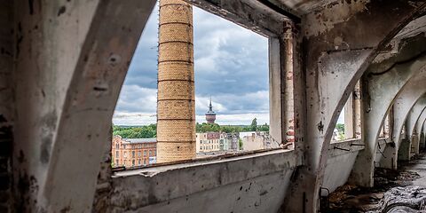 Durch das Fenster einer alten Fabrik ist ein großer Schornstein zu sehen.