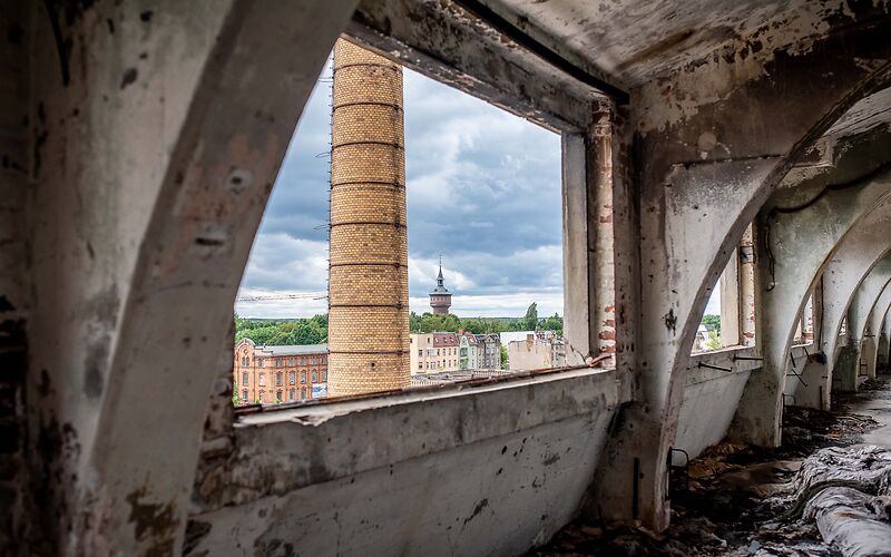 Durch das Fenster einer alten Fabrik ist ein großer Schornstein zu sehen.