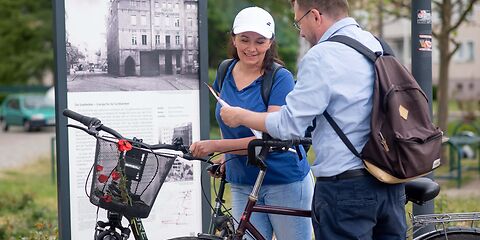 Zwei Personen, eine Frau und ein Mann, stehen mit ihren Fahrrädern vor dem Tafeln des stadtgeschichtlichen Bildungspfads Industriekultur.