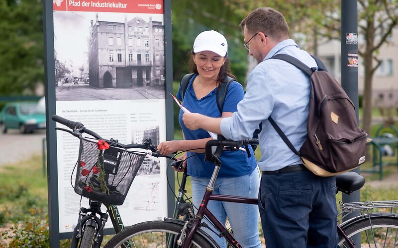 Zwei Personen, eine Frau und ein Mann, stehen mit ihren Fahrrädern vor dem Tafeln des stadtgeschichtlichen Bildungspfads Industriekultur.