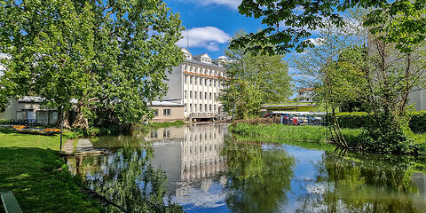 Historische Stadtmühle mit weißer Fassade am Wasser, umgeben von viel Grün, bei strahlender Sonne.
