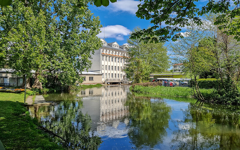 Historische Stadtmühle mit weißer Fassade am Wasser, umgeben von viel Grün, bei strahlender Sonne.
