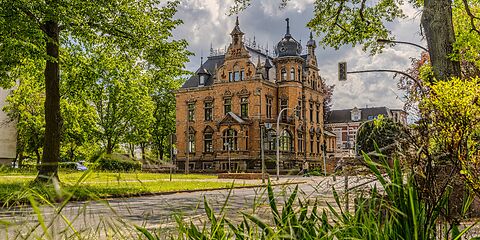 Eine große, orangefarbene Fabrikantenvilla mit dunklem Dach und mehreren kleinen Türmen. Das reich verzierte Gebäude im Neorenaissance-Stil stammt aus dem Jahr 1893. Es steht an einer Ampelkreuzung und ist von viel Grün umgeben.