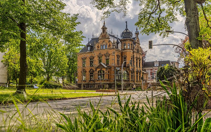 Eine große, orangefarbene Fabrikantenvilla mit dunklem Dach und mehreren kleinen Türmen. Das reich verzierte Gebäude im Neorenaissance-Stil stammt aus dem Jahr 1893. Es steht an einer Ampelkreuzung und ist von viel Grün umgeben.